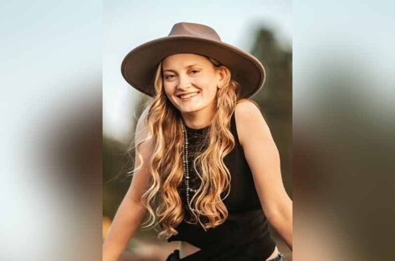 A young woman with long, wavy blonde hair smiles radiantly while leaning forward toward the camera. She is wearing a wide-brimmed tan felt hat, a black sleeveless top, and multiple long beaded necklaces. The background is a soft-focus natural landscape during the 