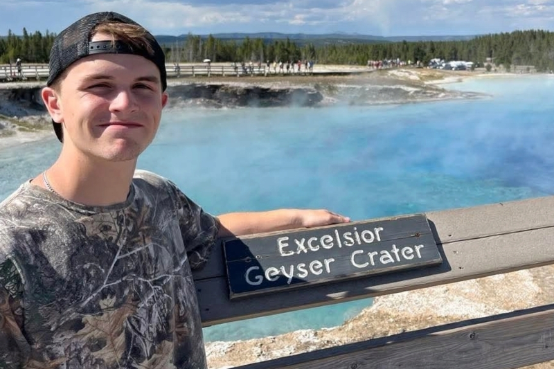 A young man in a camouflage t-shirt and backward cap smiles on a wooden boardwalk at Yellowstone National Park. He leans against a railing with a sign reading 