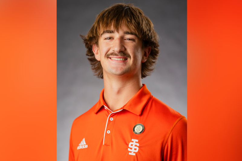 A young man with shaggy brown hair and a mustache smiles for a portrait. He is wearing a bright orange Adidas polo shirt with an 