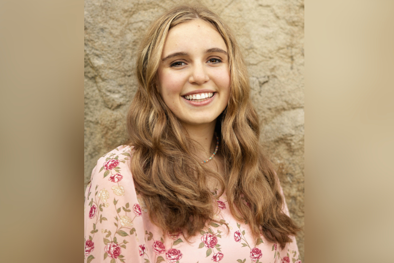 A close-up portrait of a young woman with long, wavy light brown hair and a bright, joyful smile. She is wearing a pink blouse featuring a delicate pattern of reddish-pink and cream-colored roses with green stems. She is also wearing a colorful beaded necklace. The background is a textured, light brown stone wall in soft focus.