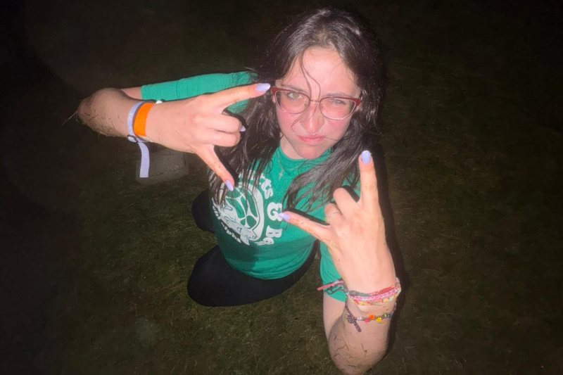 A high-angle, nighttime photo of a young woman with dark hair and glasses looking up at the camera with an intense, playful expression. She is wearing a green t-shirt and multiple colorful wristbands, holding up 