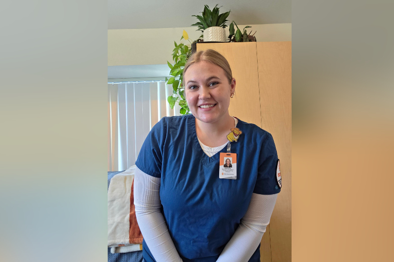 A young woman with blonde hair tied back smiles warmly in an indoor setting. She is wearing blue medical scrubs over a white long-sleeved shirt, with an orange ID badge clipped to her collar identifying her as a nursing student. A small, yellow cartoon character pin is attached to her scrubs. The background includes a wooden cabinet topped with green plants and a window with white blinds.