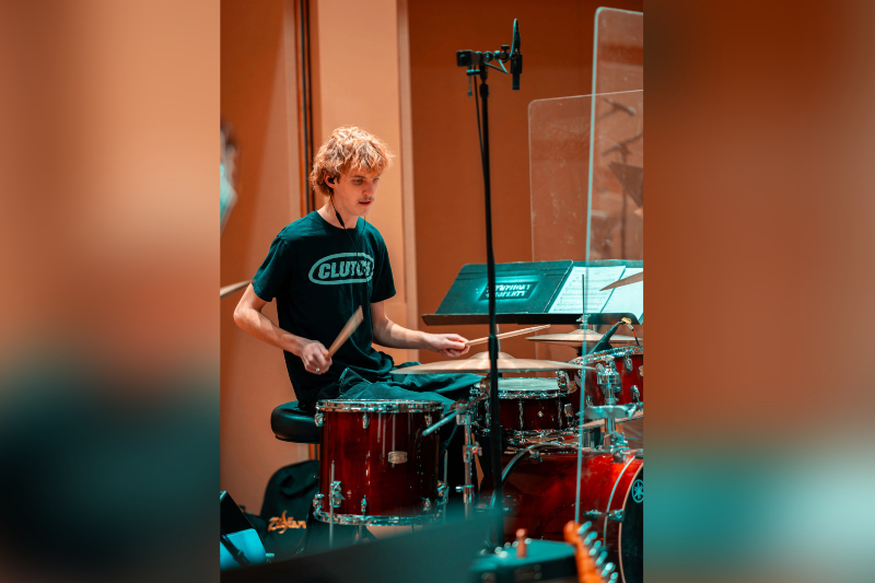 A young man with wavy ginger hair and a mustache is captured in motion playing a drum kit. He is wearing a dark t-shirt with the word 
