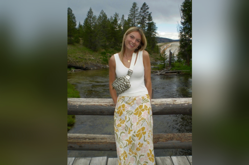 A young woman with blonde hair and a joyful smile leans slightly to one side while posing on a rustic wooden bridge. She is wearing a white ribbed tank top, a patterned skirt with yellow and pink floral designs, and a checkered cross-body bag. The background features a winding river, lush evergreen trees, and a distant view of a steaming geothermal feature or waterfall under a cloudy sky.
