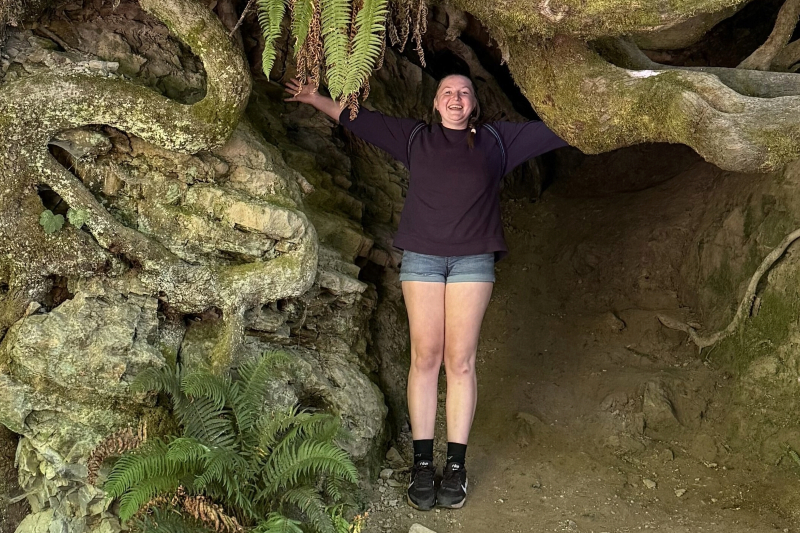 A young woman with a joyful, wide-mouthed laugh stands inside a natural cave-like opening formed by large, moss-covered tree roots and rocky earth. She has her arms outstretched to touch the sides of the cave. She is wearing a dark purple long-sleeved shirt, denim shorts, black socks, and black sneakers. Green ferns hang from the top of the cave and grow near her feet.