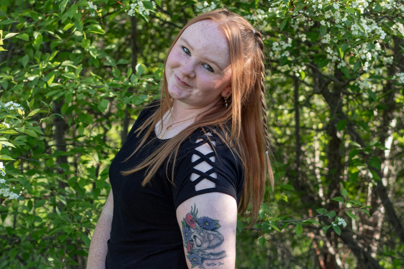 A young woman with red hair and freckles smiles while standing in front of lush green bushes with small white flowers. She is wearing a black t-shirt with criss-cross lattice detailing on the sleeves. A prominent tattoo on her upper arm features a dinosaur skull surrounded by colorful purple, blue, and red flowers. She has a small braid in her hair and is wearing a delicate gold necklace.