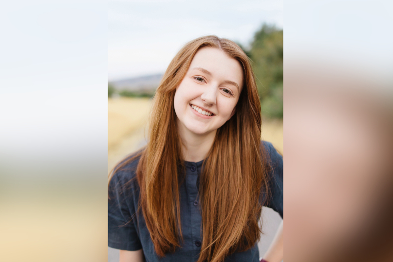 A young woman with long, straight strawberry-blonde hair smiles warmly while tilting her head slightly to the side. She is wearing a dark navy blue button-down shirt. The photo is a close-up portrait taken outdoors with a soft-focus background of golden fields and green trees under a bright, overcast sky.