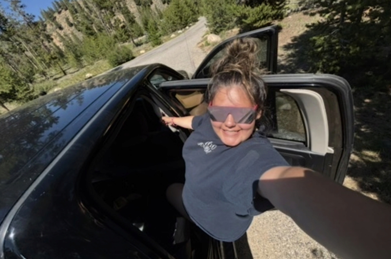 A high-angle selfie of a young woman with a messy bun and large, mirrored wrap-around sunglasses. She is smiling brightly while leaning out from a black vehicle on a sunlit mountain road. She is wearing a navy blue t-shirt. The background shows a winding road and steep, pine-covered hillsides under a clear sky.