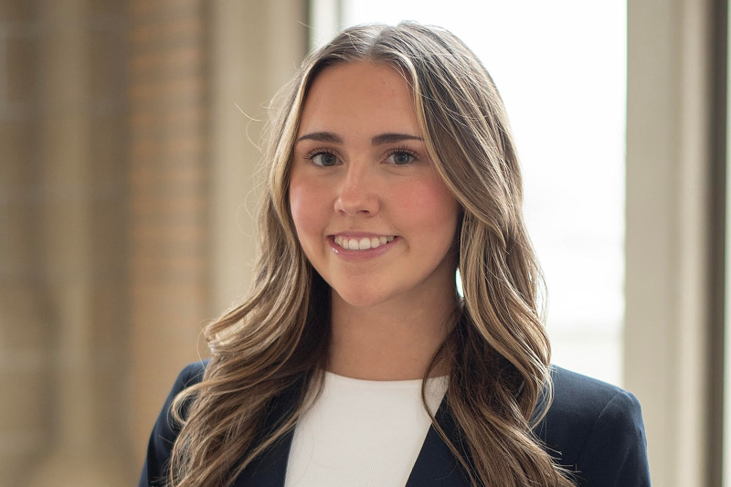 A young woman with long, wavy blonde-highlighted hair smiles warmly for a professional headshot. She is wearing a navy blue blazer over a simple white top. The background is a brightly lit indoor space with large windows and neutral-toned architecture in a soft focus.
