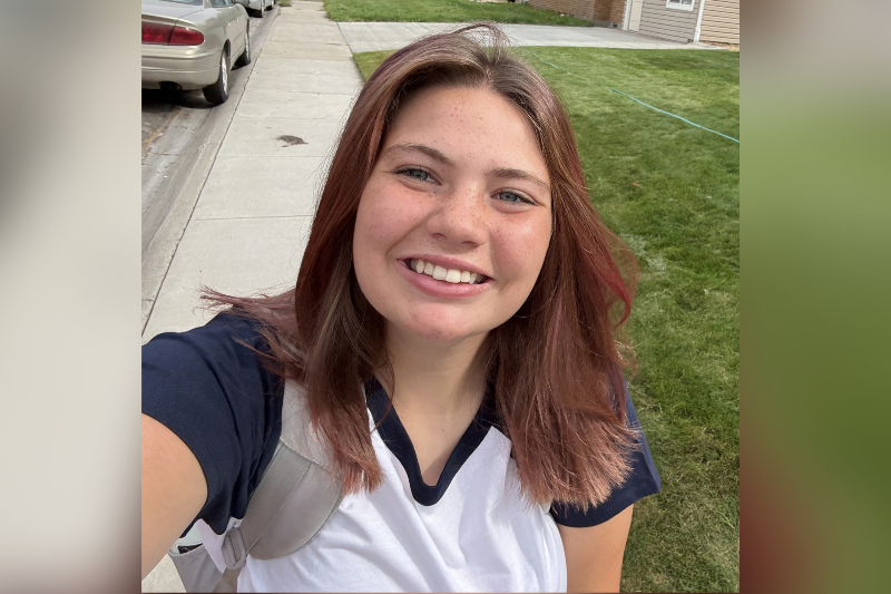 A young woman with shoulder-length reddish-brown hair and freckles smiles broadly in an outdoor selfie. She is wearing a dark blue and white ringer-style t-shirt and carrying a light grey backpack. The background shows a residential sidewalk, a parked car, and a green lawn under bright, natural daylight.
