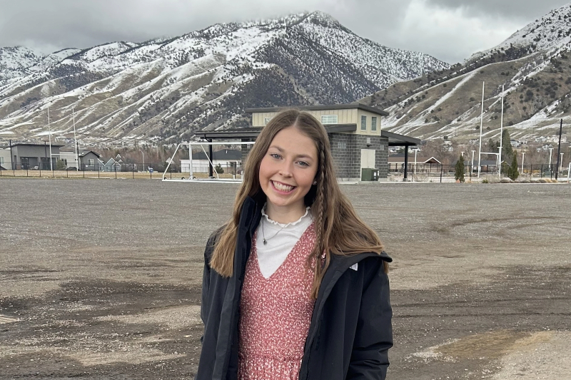 A young woman with long brown hair and a small accent braid smiles warmly for a portrait. She is wearing a pink patterned dress layered over a white ruffled top and a black jacket. She stands in a large, open gravel area with a backdrop of dramatic, snow-dusted mountains under a cloudy sky. A soccer goal and small building are visible in the distance.