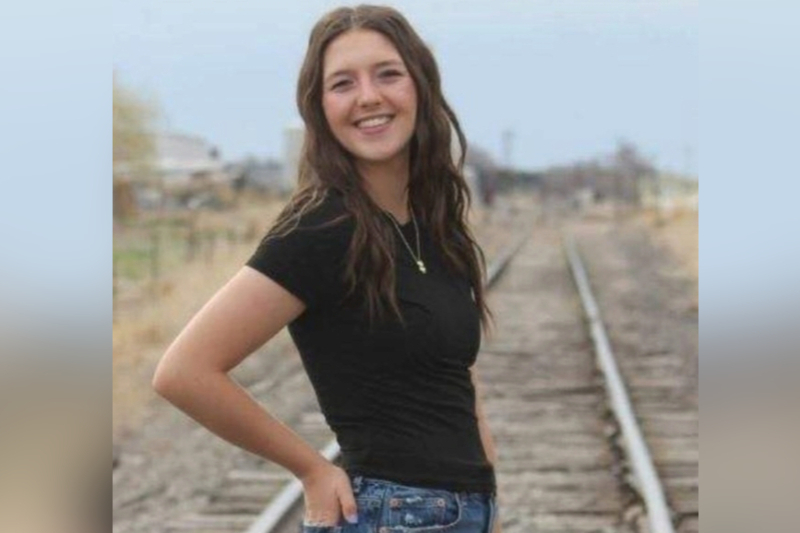 A young woman with long, wavy brown hair smiles brightly while posing on a set of railroad tracks. She is wearing a simple black t-shirt and blue jeans, with one hand tucked into her pocket. The background shows the tracks receding into the distance under an overcast sky in a rural or industrial setting.