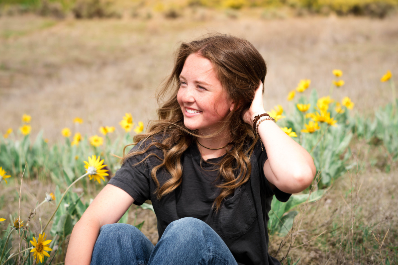 A young woman with wavy brown hair and freckles sits in a field, smiling as she looks off to the side. She is wearing a dark grey t-shirt and blue jeans, with her hand tucked behind her head. She is surrounded by bright yellow wildflowers under soft, natural light, with a blurred grassy hillside in the background.